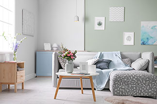 In einem Wohnzimmer steht eine grauen Eckcouch vor der ein runder Tisch mit einer weißen Tischplatte und Beinen aus hellem Holz steht.