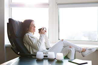 Relaxen im Sessel: Eine weiß angezogene Frau sitzt vor einer Fensterfront in einem schwarzen Relaxsessel aus Leder und hält eine Tasse Tee in der Hand.