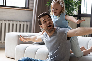 Geprüfte Qualität im Wohnzimmer: Ein Vater sitzt mit ausgestreckten Armen vor der Couch und spielt mit seiner kleinen Tochter, die hinter seinem Rücken auf dem Sofa kniet und die Arme ausbreitet.