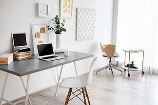 Der richtige Arbeitsplatz: Ein grauer Schreibtisch auf dem ein Laptop steht, befindet sich an einer weißen Wand an der zwei Dekobilder und eine Uhr hängen – davor ein weißer Stuhl.