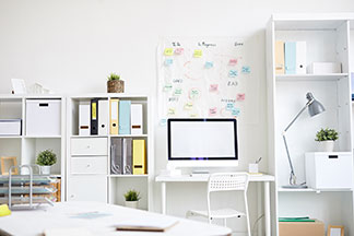 Ordnung am Schreibtisch: Ein weiß eingerichtetes Arbeitszimmer mit einem Schreibtisch, der zwischen zwei Regalen steht, in denen Ordnern und Aufbewahrungsboxen stehen.