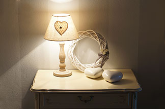 Ein rechteckiger weißer Nachttisch mit Schublade, auf dem sich zwei Keramikherzen, ein geflochtener Holzkranz sowie eine Lampe befinden, steht im Flur vor einer weißen Wand.