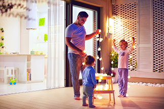 Im Dämmerlicht stehen ein Vater, sein kleiner Sohn und seine kleine Tochter auf der Terrasse, und beschäftigen sich mit einer leuchtenden Lichterkette mit kleinen, runden Lampen.