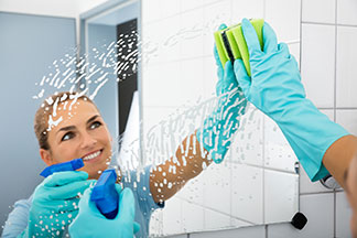 Badspiegel richtig reinigen: Eine junge Frau putz mit blauen Handschuhen und einem Reinigungsspray ihren Spiegel.