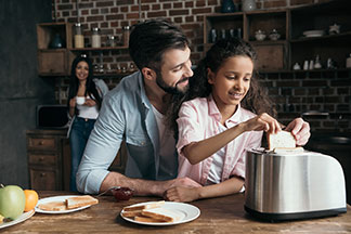 Gemeinsamen Frühstück: Ein junger Vater steckt zwei Toastscheiben in einen silbernen Toaster, der auf der Küchenanrichte steht.