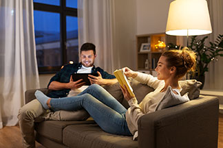 Entspannung im Wohnzimmer: Ein junges Paar liegt am Abend gemütlich mit einem Buch und einem Tablet auf der Wohnzimmercouch.