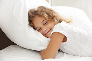 Gute Qualität für guten Schlaf: ein kleines Mädchen liegt auf dem Bauch in ihrem weiß überzogenen Bett und kuschelt sich in ihr Kopfkissen.