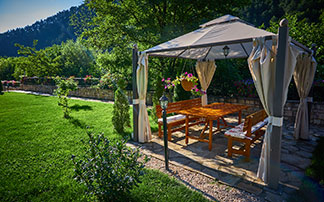 In einem grünen Garten steht ein Pavillon mit einem Aluminiumgestell, einem weißen Stoffdach sowie aufgezogenen Stoffwänden an den Seiten. Darunter steht ein rechteckiger Holztisch mit Holzbänken.
