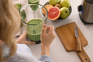 Küchengeräte im Einsatz: Eine Frau steht in der Küche und hält ihren Mixer mit einem grünen Smoothie darin in der Hand – daneben eine Schale Obst und ein Holzschneidebrett mit Messer.