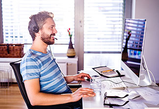 Richtige Möbel für den Arbeitsplatz: Ein junger Mann sitzt mit Headset an seinem heimischen Arbeitsplatz vor einem Monitor.