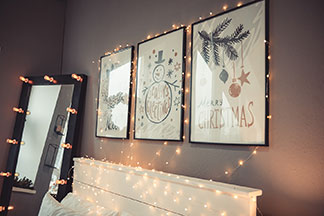 Dekoleuchten für Bilder: An einer grauen Wand und über einem weißen Bett hängen drei Bilder in schwarzen Rahmen, die mit einer Lichterkette umrandet sind.