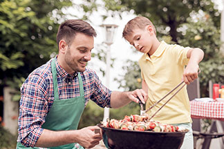 Ein junger Mann, der eine grüne Schürze trägt, kniet neben einem schwarzen, runden Grill, während sein Sohn mit einer Zange Fleischspieße wendet.