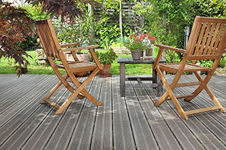 Zwei Gartenstühle aus Holz stehen auf einer Terrasse mit Blick in einen grünen Garten – vor ihnen ein kleiner viereckiger Holztisch mit einer Blumenvase darauf.ö