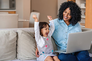 Gute Qualität lohnt sich: Eine Frau sitzt auf der Couch und hält ihren Laptop auf dem Schoß, daneben befindet sich ihre kleine Tochter, die beide Arme in die Höhe streckt und sich freut.
