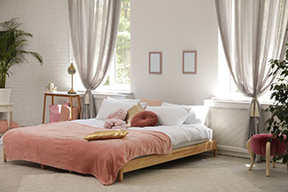 Heimtextilien für Wohlfühlatmosphäre: Ein großes Schlafzimmer mit einem schön bezogenen Doppelbett mit rosa Tagesdecke, Dekokissen und weißen Vorhängen.