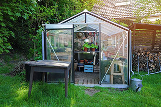 Ein Gewächshaus – mit einem Spitzdach und einer offen stehenden Tür – steht in einem grünen Garten.