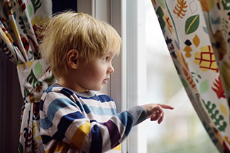 Ein kleiner Junge schaut interessiert aus einem Fenster, an dem bunte Gardinen hängen.