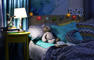 Eine junge Mutter liegt mit ihrem Kleinkind im Bett und liest ein Buch vor, während die Nachttischlampe brennt.