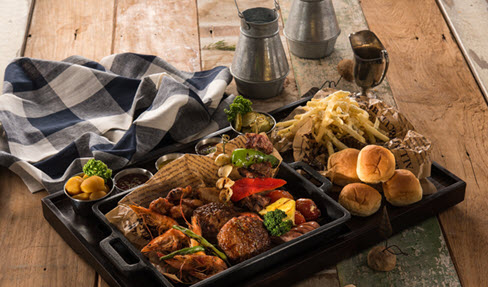 Servierplatte dekoriert mit vielen Spezialitäten vom Grill: Garnelen, Fleischspieße und Gemüse. Dazu Pommes in einer braunen Papiertüte und Pizzabrötchen.