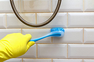 Mit den richtigen Putzutensilien strahlt das Bad: Eine Hand, die einen gelben Handschuh trägt, reinigt die Dusche mit einer blauen Fugenbürste.