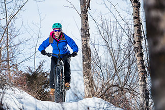 Eine junge Frau fährt auf dem Rad bergauf durch eine winterliche Landschaft und trägt winterliche Fahrradbekleidung, die aus einer blauen Jacke und schwarzen Hosen besteht.