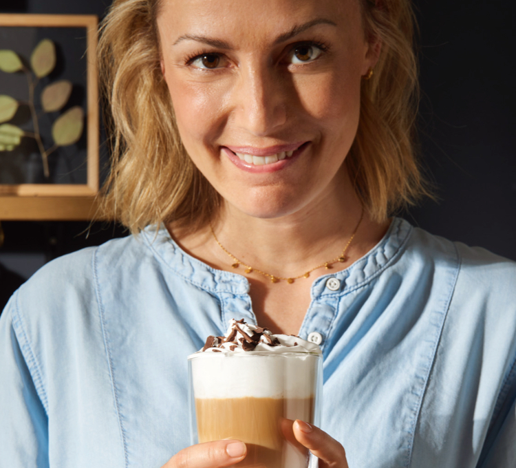 Eine lächelnde Frau trinkt einen Kaffee mit Milchschaum aus einem Latte Macchiato Glas