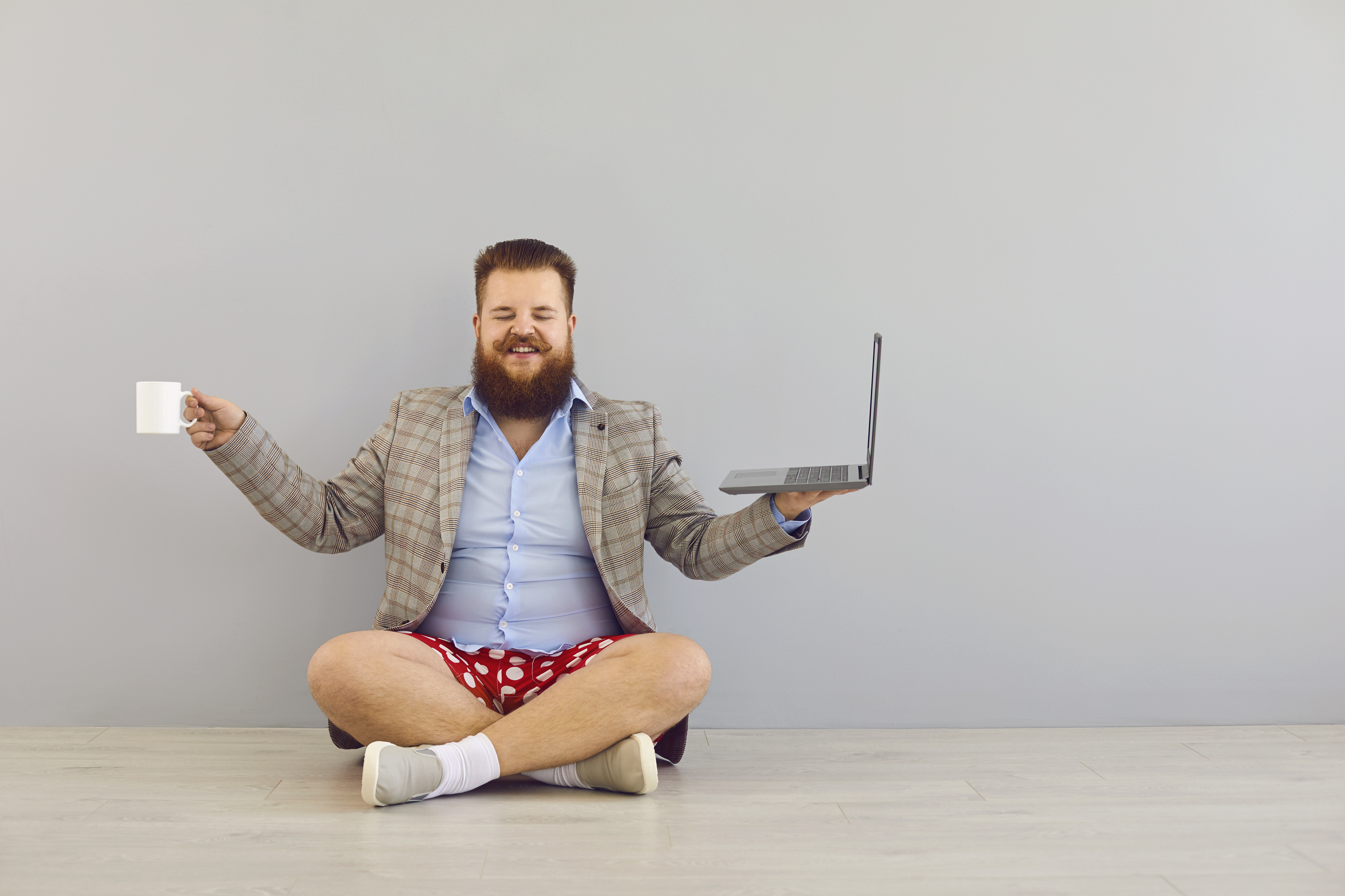 Mann mit Laptop, Kaffeetasse sowie Sakko, Hemd und Boxershorts im Schneidersitz.