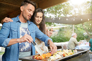 Ein junger Mann steht am Grill und wendet einen Fleischspieß mit einem Schaber, während eine junge, langhaarige Frau ihren Arm um seine Schulter legt, und ebenfalls auf den Grill schaut.