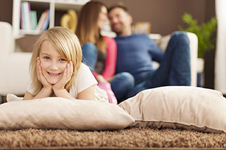 Qualität, die passt: Ein junges Mädchen liegt gemütlich auf zwei nebeneinander liegenden Kissen auf dem Wohnzimmerboden, während sich die Eltern auf der Couch im Hintergrund unterhalten.