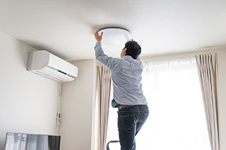 Deckenlampe für niedrige Räume: Ein Mann in blauer Jeans und Hemd steht auf einer Leiter, und ist gerade dabei eine Tellerlampe an der Decke zu montieren.