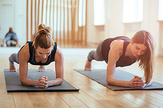 Zwei Frauen, die beide eng anliegende Sporthosen tragen, befinden sich im Unterarmstütz auf zwei Yoga-Matten.