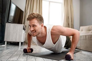 Ein junger Mann bei seinen Fitnessübungen mit Hanteln im heimischen Wohnzimmer auf einer Fitnessmatte.