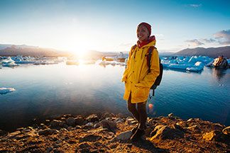 Eine junge Frau, die mit einem gelben Regenmantel und einer roten Mütze bekleidet ist, steht bei Sonnenschein in Island vor einem Gewässer, auf dem Eisberge schwimmen.
