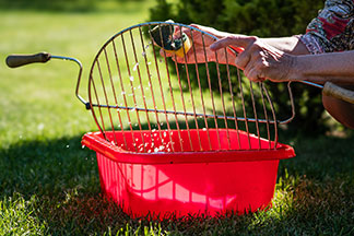 Grill richtig reinigen: Zwei Hände reinigen einen Grillrost in einer roten viereckigen Plastikschüssel mit Wasser.