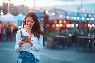 Gut erreichbar: Eine junge Frau bedient draußen ihr Smartphone, während im Hintergrund ein Straßenmarkt im Abendlicht zu sehen ist.