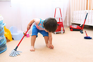 Putzutensilien für die Kleinen: Ein kleiner Junge bückt sich zum Teppich, während er den Boden seines Kinderzimmers mit extra Kinder-Putzutensilien reinigt.