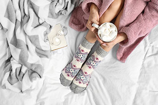 Für die kühle Jahreszeit: Eine Frau in einem rosa Bademantel, mit gemusterten dicken Strümpfen und angezogenen Beinen sitzt mit einer weißen Tasse voller Marshmallow auf ihrem Bettlaken.