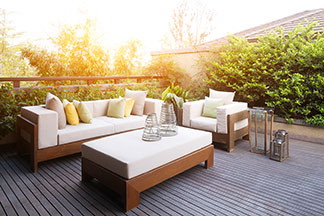Vor einer offenen Terrassentür stehen Loungemöbel aus geflochtenem Polyrattan sowie zwei Sitzpolster. Der Tisch ist gedeckt und auf der Couch liegen mehrere Dekokissen.
