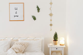 Lampen in Tchibo-Qualität: Ein weiß eingerichtetes Schlafzimmer mit einem Bett und einem Nachttisch, auf dem eine angeschaltete, pilzförmige Dekoleuchte steht.