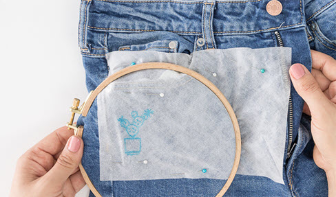 Ein bemaltes Vlies wird auf einer Jeanshose mit Stecknadeln befestigt