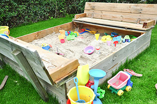 In und neben einem Sandkasten aus Holz, der auf einer Wiese steht, liegt buntes Spielzeug für draußen – darunter Sandförmchen, Schaufeln und Eimer.