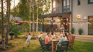 Gute Qualität für Ihre Gartenstühle: In einem Garten sitzen fünf Erwachsene am Tisch beim Essen, während zwei Kinder herumtollen. Über Ihnen hängt eine angeschaltete Lampionkette.