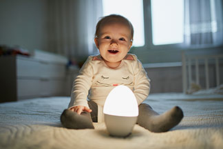 Sicherheit durch Qualität: Ein Baby sitzt lachend auf einem Bett vor einer eiförmigen Lampe.