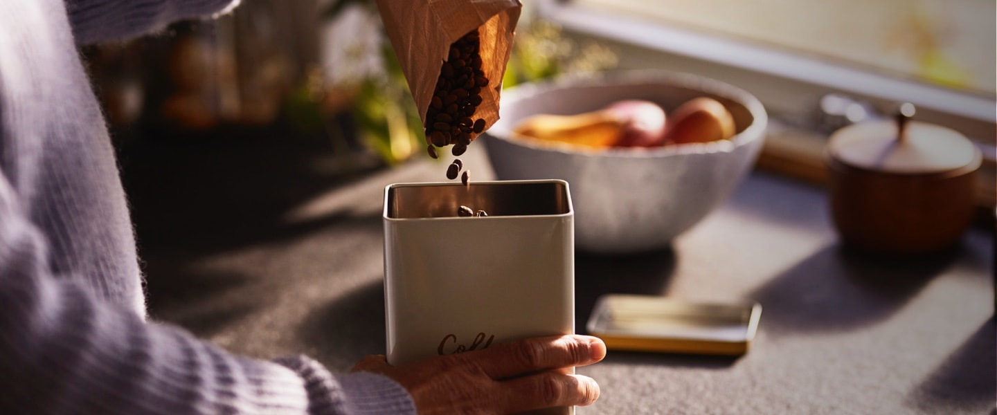 Kaffeebohnen werden in eine Kaffeedose verkippt. Dieses stimmungsvolle Kaffeegenussbild leitet den Schwerpunkt der Kaffeeaufbewahrung ein.