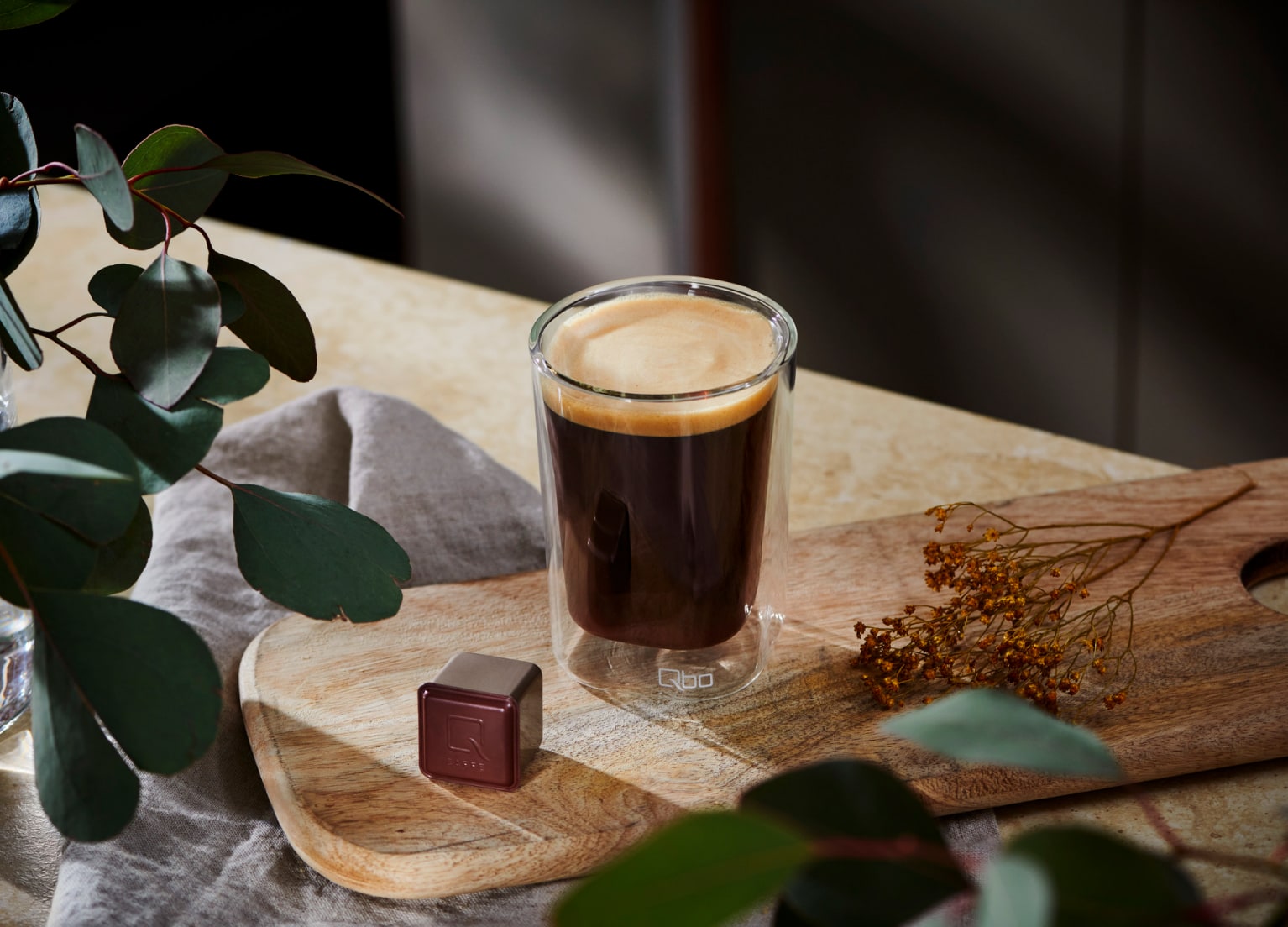 Ein Glas mit Kaffee und eine Qbo Kapsel liegern auf einer Holzplatte