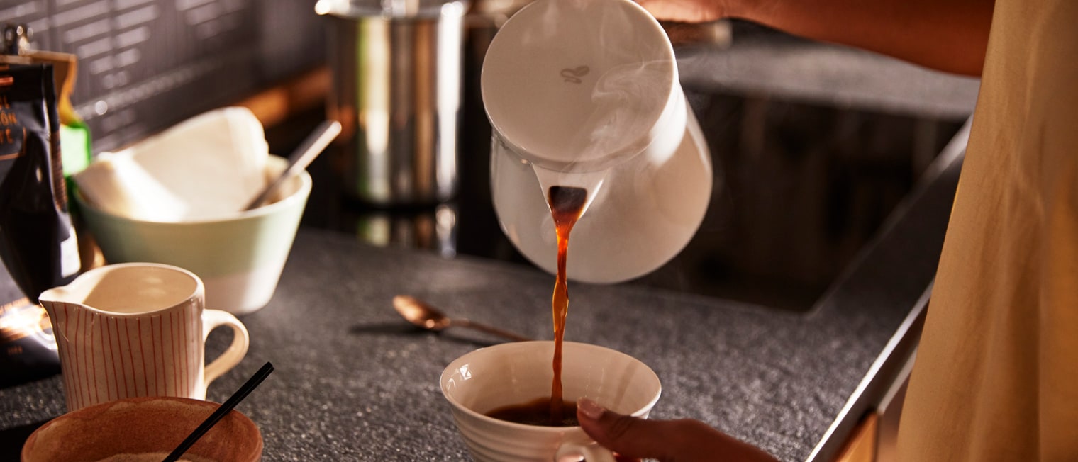 Filterkaffee wird auf einer Küchentheke aus einer Porzellankanne in eine Kaffeetasse eingegossen.