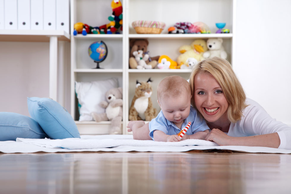 Eine blonde, lachende Frau liegt mit ihrem Baby, das mit einem rot-weiß-gestreiften Spielzeug spielt, auf einer Decke auf dem Boden im Kinderzimmer.