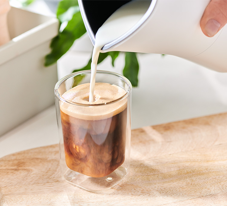 Milchschaum wird aus einem Milchaufschäumer in ein Caffè Crema Glas gegossen.