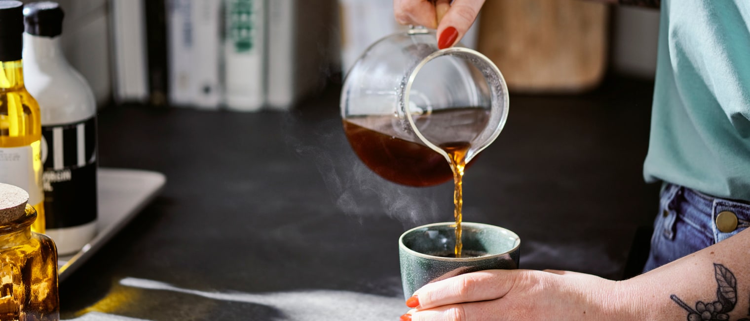 Eine Person steht an einer Küchentheke und schenkt sich Specialty Coffee Filterkaffee aus einer Glaskanne in einen Kaffeebecher ein.