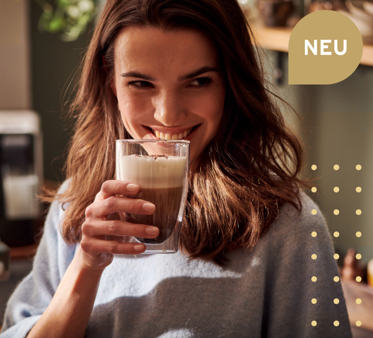 Eine Frau trinkt eine Kaffeespezialität mit Milch und lächelt dabei